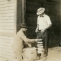 Black and white photograph of two individuals tying up ears of corn on a rack for drying, c.1910