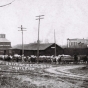 Black and white photograph of horse-drawn wagons hauling bags of seed corn to a railroad station for further transporting, c.1907.