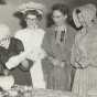 Black and white photograph of Mary Walker, a BPW charter member, lighting a kerosene lamp, 1958. With her are (left to right) Mary Walker, Mae Rohrer, Elizabeth Marsh, and Sylvia Johnson, all wearing historic costumes.