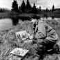 Birney Quick painting a landscape out in nature, along the shore of a lake, with pine trees in the background.