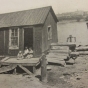 Black and white photograph of a shack and outhouse, river’s edge, 1917