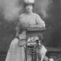 Photograph of Mrs. Emile Amblard sitting on a wicker chair with a tennis racquet in ther hand, a boat cap on her head, and a dog seated by her side. Circa 1910..