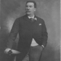 Photograph of Emile Amblard leaning on a table, holding his gloves and wearing a boutonniere. Circa 1910.