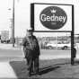Black and white photograph of a man standing to the left of the Gedney sign, outside the Chaska factory,  c.1980s.