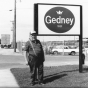 Black and white photograph of a man standing to the left of the Gedney sign, outside the Chaska factory,  c.1980s.