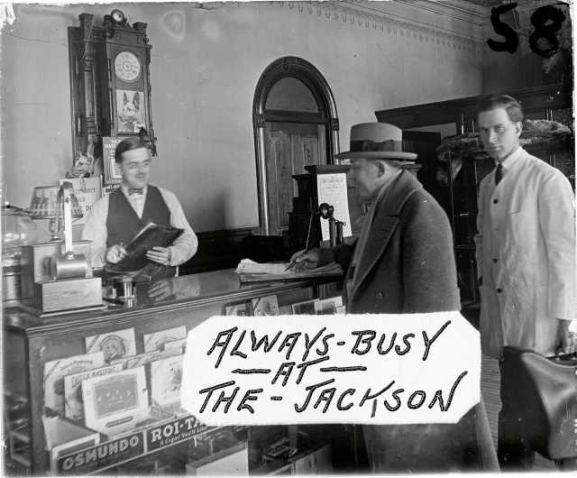 Image from a glass plate negative showing the front desk of the Jackson Hotel, ca. 1926. Cigars for sale are visible in the case, and a timetable for the Anoka Electric Line is fixed to the back of the cash register. Art Anderson is one of the men in the photograph. This image was used as an advertisement at Green’s Theater, located on Main Street in Anoka a few blocks from the hotel. Photographer unknown. Used with the permission of the Anoka County 