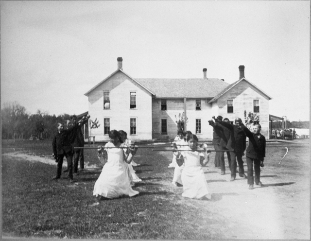 Black and white photograph of students performing a drill on the grounds of a Native American boarding school, c.1890s.