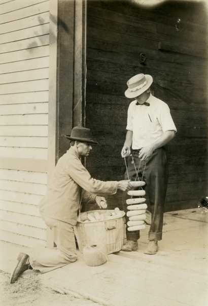 Black and white photograph of two individuals tying up ears of corn on a rack for drying, c.1910