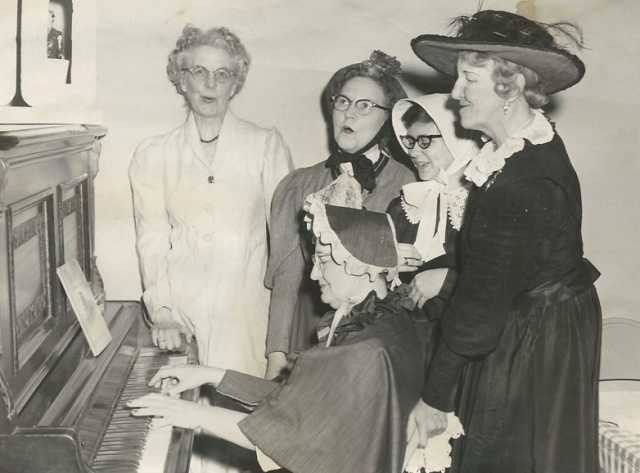 Black and white photograph of BPW members in historic costumes singing around a piano, 1958. Pictured (left to right) are Louise Rasmussen, Clara Berg, Ruth Rohrer (piano player), an unknown woman, and Maybelle Anderson.