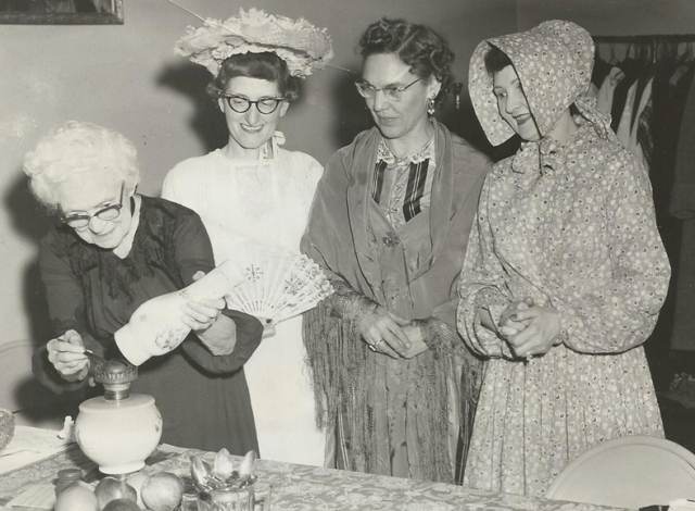 Black and white photograph of Mary Walker, a BPW charter member, lighting a kerosene lamp, 1958. With her are (left to right) Mary Walker, Mae Rohrer, Elizabeth Marsh, and Sylvia Johnson, all wearing historic costumes.