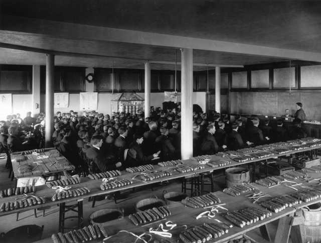 Black and white photograph of a large lecture room at University of Minnesota with ears of corn laid out for judging by students. Photographed by Harry D. Ayer c.1910.