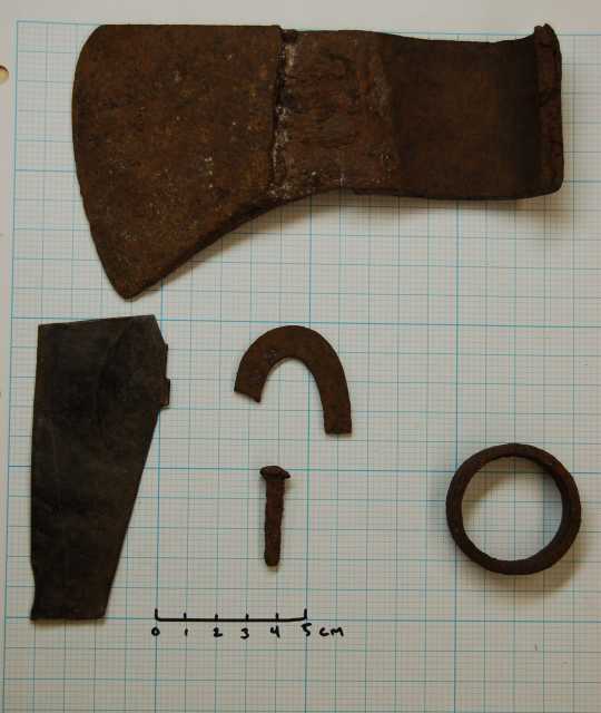 Color image of artifacts metal detected by a relic collector prior to 1972 and identified as an iron ferule, a piece of fire steel, a rosehead nail, a fragment from the bottom of a kettle, and an axe head.
