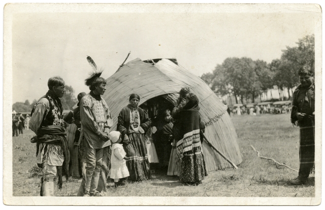 An Ojibwe family standing by bull rush wigwam