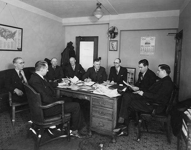 photograph of a group of men gathered around a desk in the State Office Building