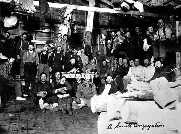 Photograph of a small congregation of lumberjacks assembled inside a bunkhouse to hear sky pilot Frank Higgins preach c.1910.