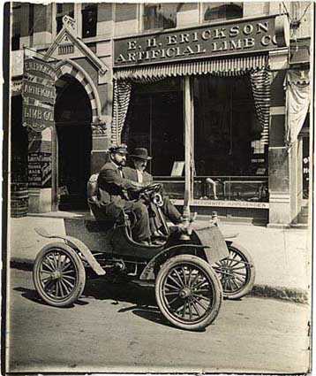 Black and white photograph of Michael Dowling and a passenger in a car in front of the E.H. Erickson Artificial Limb Company, c.1905.