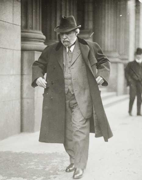 Black and white photograph of James J. Hill walking down street with overcoat flapping, c.1915.