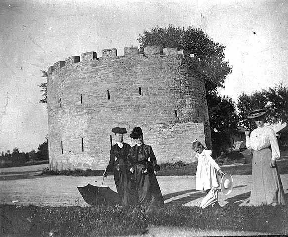 Black and white photograph of women with parasols by round tower, c.1900.