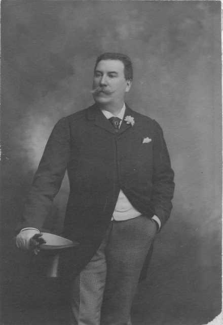 Photograph of Emile Amblard leaning on a table, holding his gloves and wearing a boutonniere. Circa 1910.
