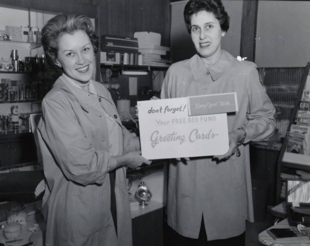 Black and white photograph of two members of the Mount Sinai Women's Auxiliary posing in the Mount Sinai Hospital gift shop holding a sign advertising greeting cards, c.1954.