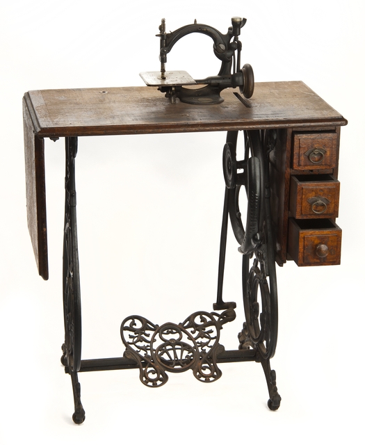 Noiseless Automatic Sewing Machine designed by by the Willcox & Gibbs Sewing Machine Company in London, England. The treadle and gears run smoothly on this chain stitch sewing machine. The machine is mounted on a wooden table with an iron base, a foot treadle, and three drawers.