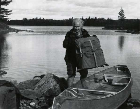 Black and white photograph of Dorothy Molter lifting a pack out of a canoe, Isle of Pines, Knife Lake, Boundary Waters Canoe Area, ca. 1960s.
