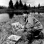 Birney Quick painting a landscape out in nature, along the shore of a lake, with pine trees in the background.