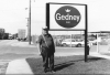 Black and white photograph of a man standing to the left of the Gedney sign, outside the Chaska factory,  c.1980s.