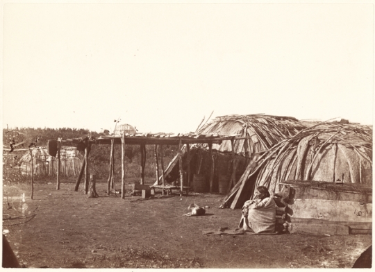 Black and white photograph of a Ho-Chunk woman sitting outside a shelter. Taken by Benjamin Franklin Upton in 1858.