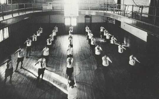 Black and white photograph of girls in uniform performing calisthenics during a physical education class on the second floor of Kiehle Hall, 1927. 