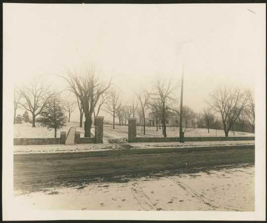 The stone fence added to the Woodbury House property by the Caswell family in 1911. Both the vehicle and pedestrian gates are visible, opening onto an unpaved Ferry Street. Photographer and exact date unknown. Anoka County Historical Society, Object ID# 2074.1.5. Used with the permission of Anoka County Historical Society.