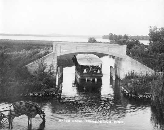 photograph of small steamboat passing under a bridge