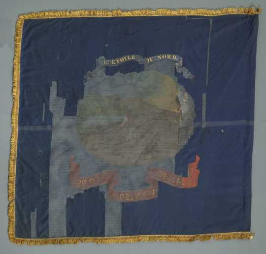 The Fourth Minnesota battle flag is blue with gold fringe. It is missing a large portion of the center, but a motto and image are still somewhat visible. This is the reverse side.