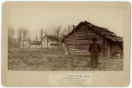 photograph shows Peterson in front of log house, with newer house in background