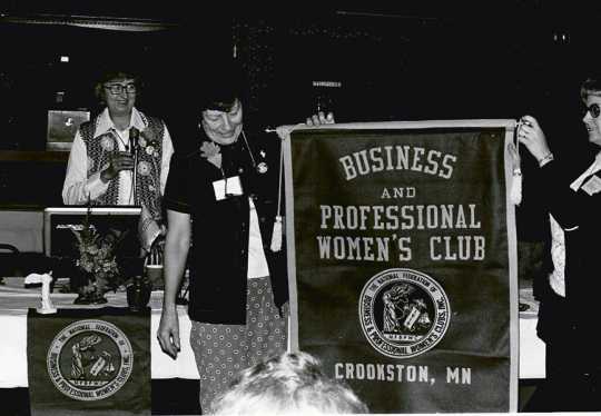 Black and white photograph of Crookston BPW president Betty Brecto watches as BPWC members Eleanor Johnson and Norma Van Horn, two BPW members, reveal the new Crookston BPWC banner on March 11, 1977.