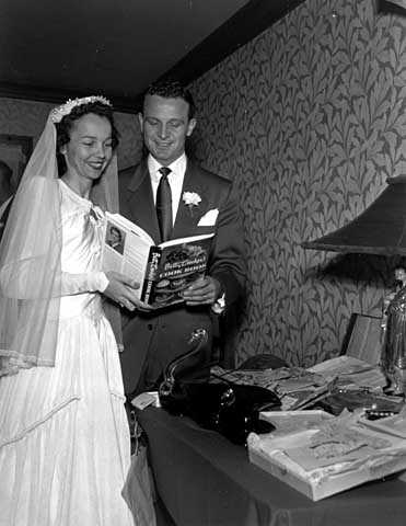 Black and white photograph of a wedding in the family of Mr. and Mrs. Frank Uram; bride and groom at gift table looking at Betty Crocker cook book, 1950. 