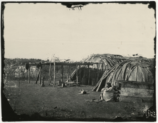 Black and white photograph of a Ho-Chunk woman sitting outside a shelter. Taken by Benjamin Franklin Upton in 1858.