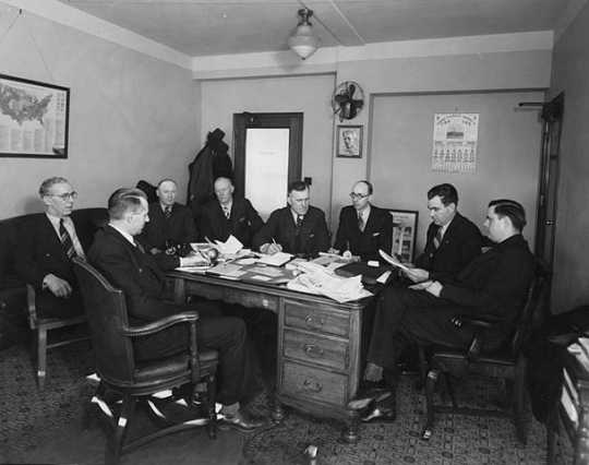 photograph of a group of men gathered around a desk in the State Office Building