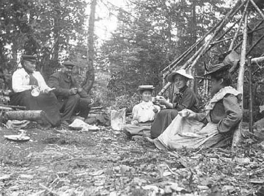 Black and white photograph of Frances Densmore seated on ground with others at Pigeon River, c. 1905.