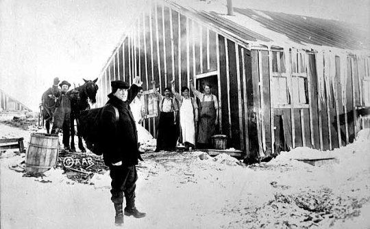 Photograph of men waving to Frank Higgins, the lumberjack sky pilot, outside a lumber camp c.1910.