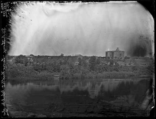 Black and white photograph of St. Anthony looking across Nicollet Island at Winslow House, right, and Universalist Church, left, ca. 1860. 