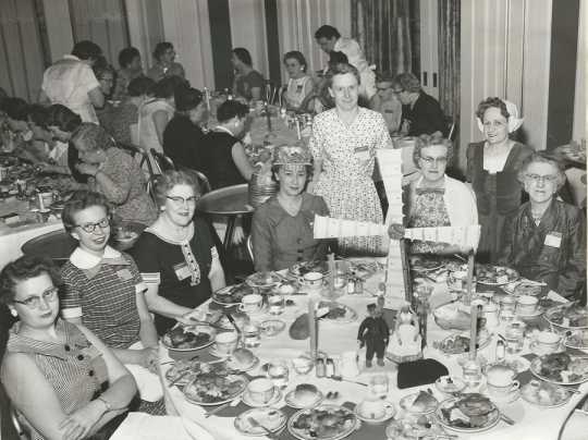 Black and white photograph of Crookston BPW club members at a table representing Denmark during an international breakfast event, 1958.
