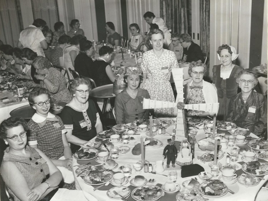 Black and white photograph of Crookston BPW club members at a table representing Denmark during an international breakfast event, 1958.