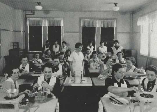 Black and white photograph of Retta Bede with students during a home-cooking class, 1930s.