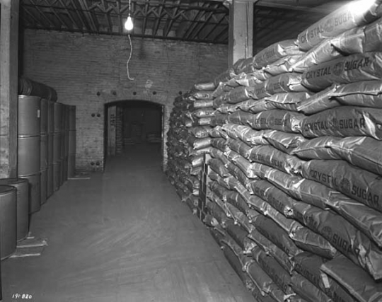 Black and white photograph of sacks of sugar produced by the American Crystal Sugar Company sit in a storage warehouse. Photographed by Norton & Peel on November 30, 1949