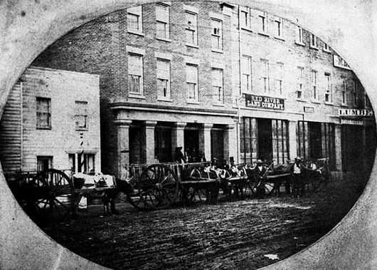 Black and white photograph of Red River Carts loading supplies at a trading house in St. Paul, 1854.