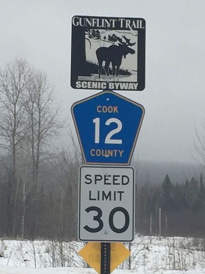 A sign assembly at the start of the Gunflint Trail indicating the Scenic Byway, county road designation, and speed limit. Photograph by Wikimedia Commons user Molandfreak, January 25, 2016. GNU free documentation license 1.2.