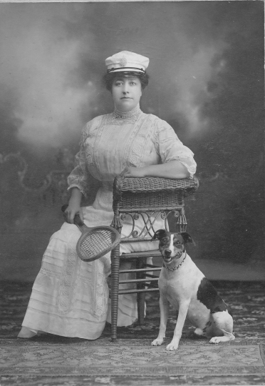 Photograph of Mrs. Emile Amblard sitting on a wicker chair with a tennis racquet in ther hand, a boat cap on her head, and a dog seated by her side. Circa 1910..