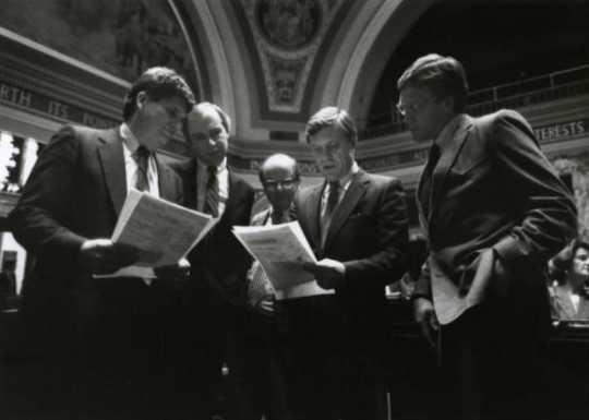 Black and white photograph of Ron Dicklich, William Luther, Allan Spear, Roger Moe, and Duane Benson reading a bill on the floor of the Minnesota Senate, 1988. 
