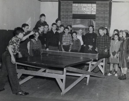 Black and white photograph of two boys playing ping pong at the Jewish Educational Center Annex while a group of children looks on, 1940.